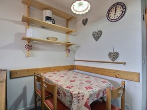 Ett litet, ombonat rum med en säng i trä, ett bord med ett mönstrat bordsduksöverdrag, hyllor med diverse föremål och dekorativa, hjärtformade väggdekorationer.