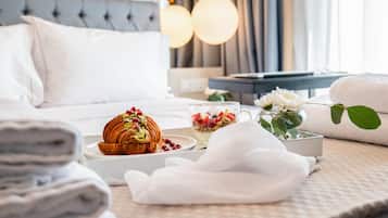 A neatly arranged hotel room with a bed, a bedside table with a lamp, a bowl of fruit, a croissant, and a vase with flowers.