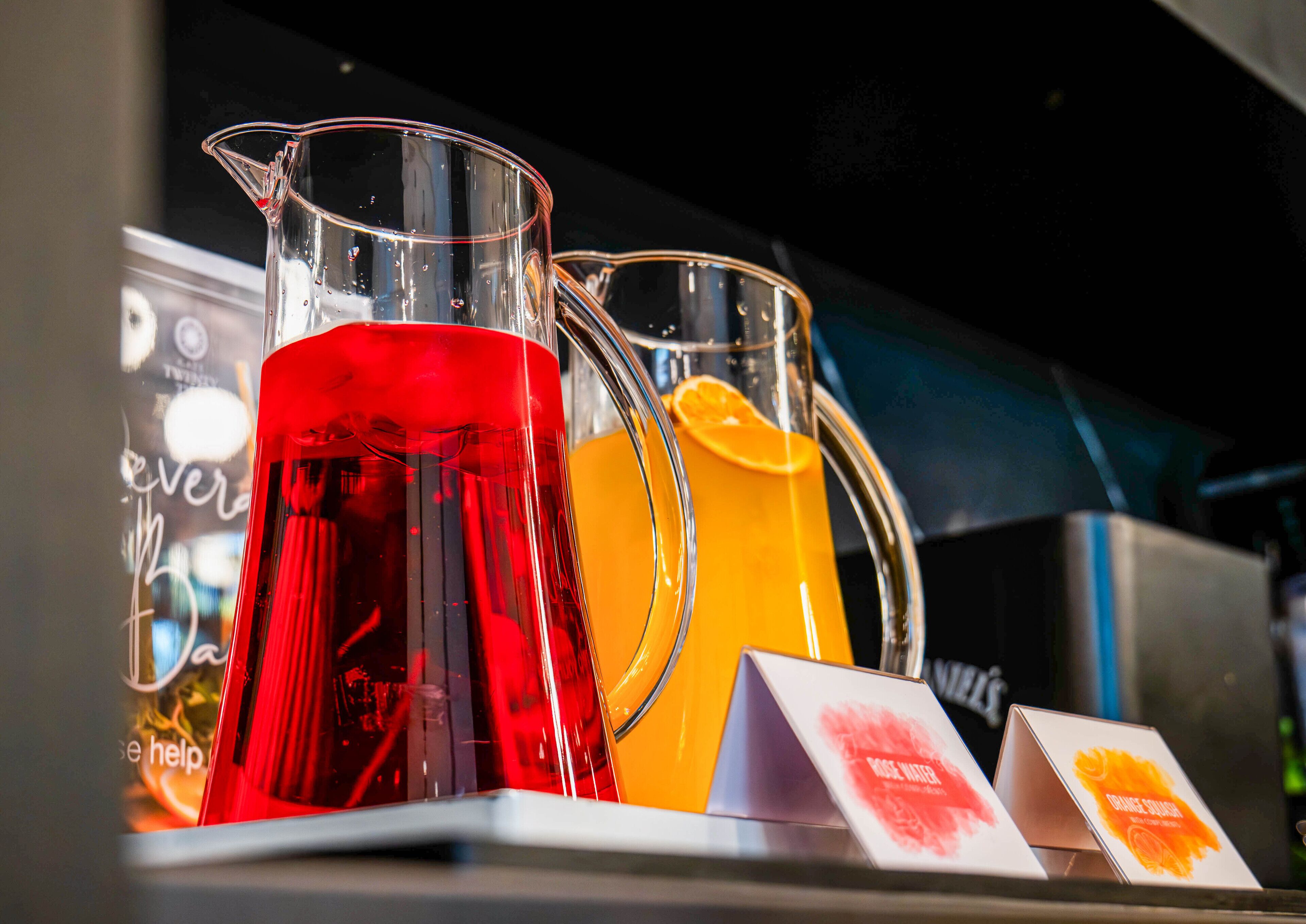 A display of pitchers with red and orange liquids, possibly juices, with labels indicating 'Rose Water' and 'Orange Juice'.