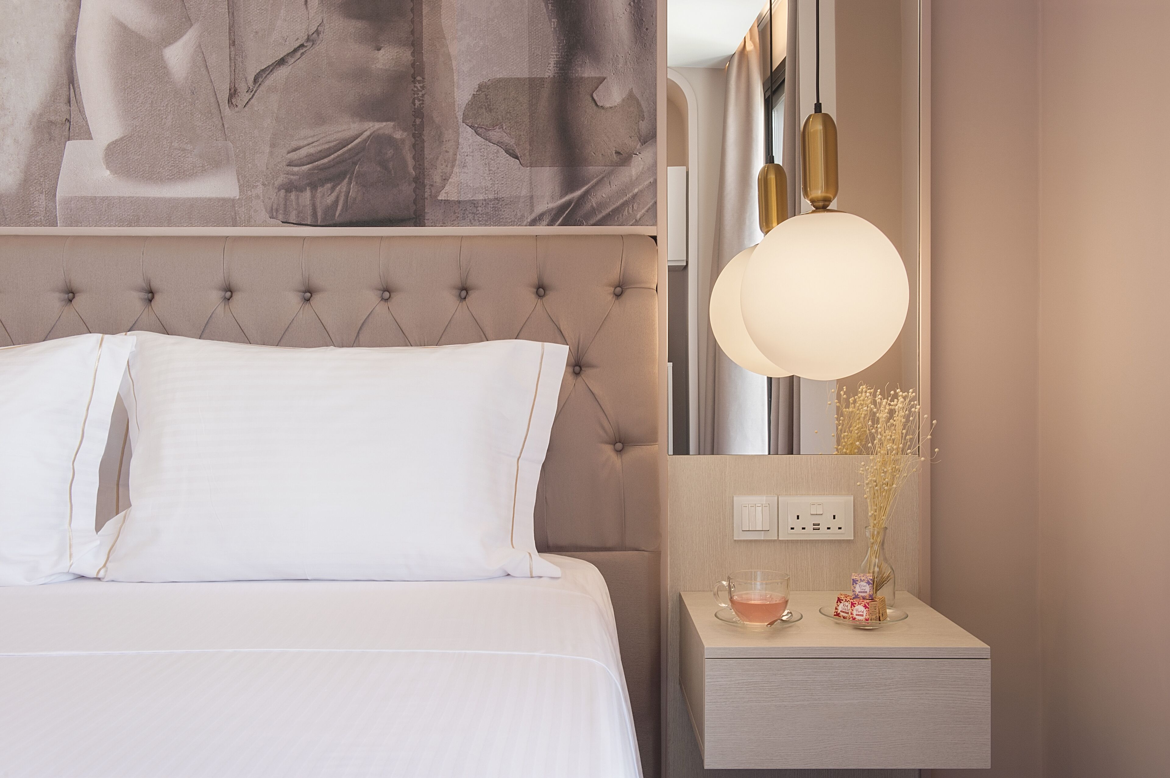 Un lit bien fait, avec du linge de lit blanc, une tête de lit capitonnée et une table de chevet sur laquelle se trouvent une lampe, un vase et une petite boîte.