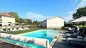 Piscina com espreguiçadeiras e guarda-sóis, rodeada por um deck de madeira e edifícios.