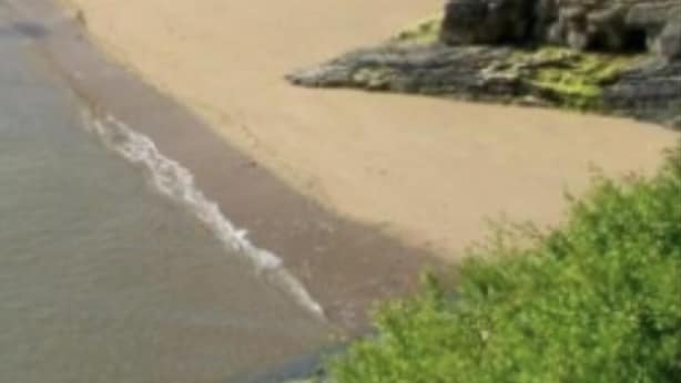Ein Strand mit einigen Personen, einer Steinmauer und Gebäuden im Hintergrund.