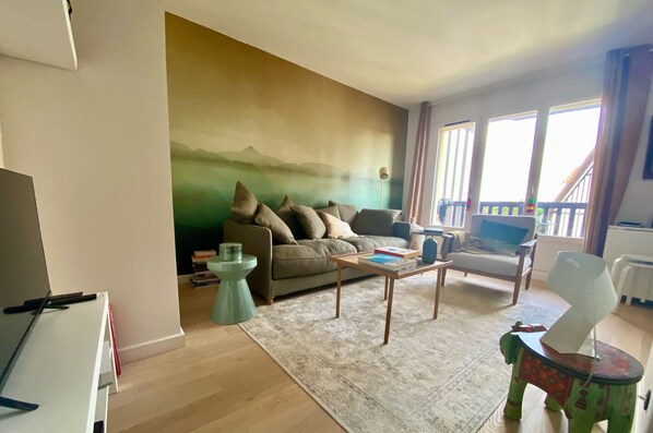 A living room with a sofa, coffee table, and a colorful stool. There is a painting on the wall and a window with curtains.
