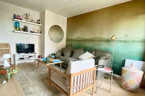 A living room with a sofa, TV, bookshelf, and a painting of mountains.