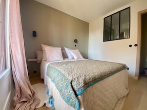 Une chambre à coucher comprenant un lit, une table de chevet, une lampe fixée au mur et une fenêtre avec des rideaux.