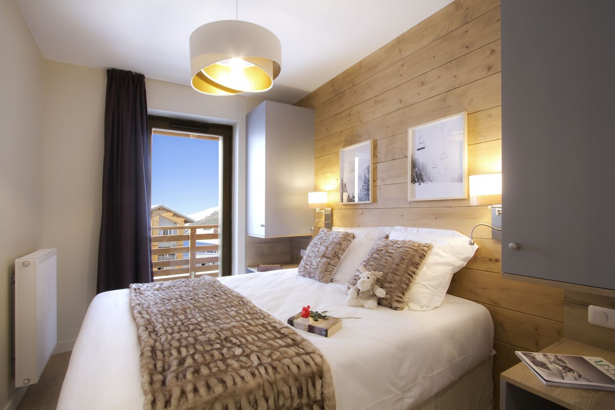 A bedroom with a bed, a bedside table, a lamp, a window with a view, and a wooden wall.