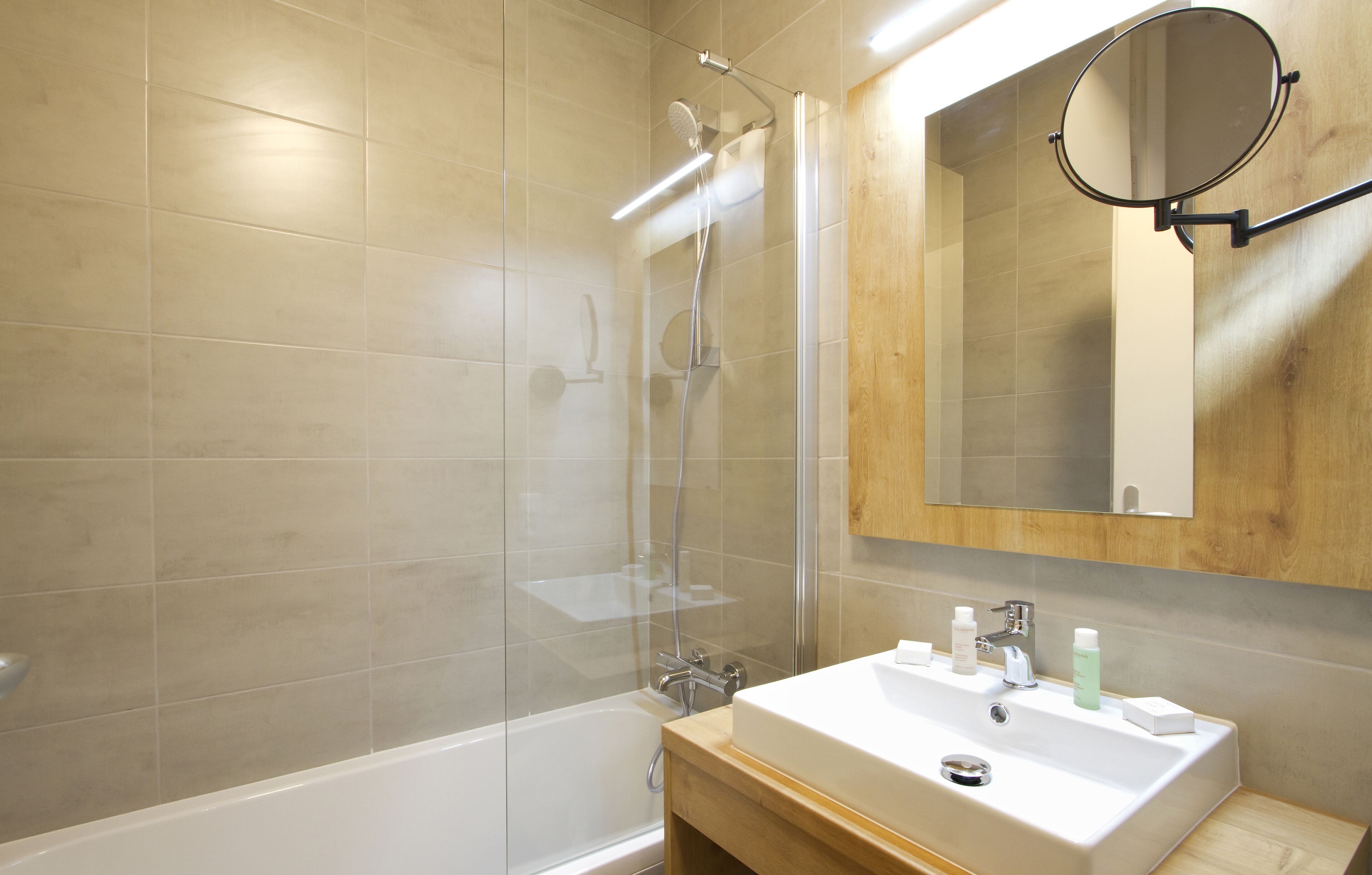 Un baño moderno con ducha envuelta en vidrio, un lavamanos blanco con artículos de aseo y un mueble de tocador de madera.