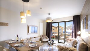 Une chambre d’hôtel moderne dotée d’un coin repas, d’un canapé et d’un balcon offrant une vue sur les bâtiments et les montagnes.