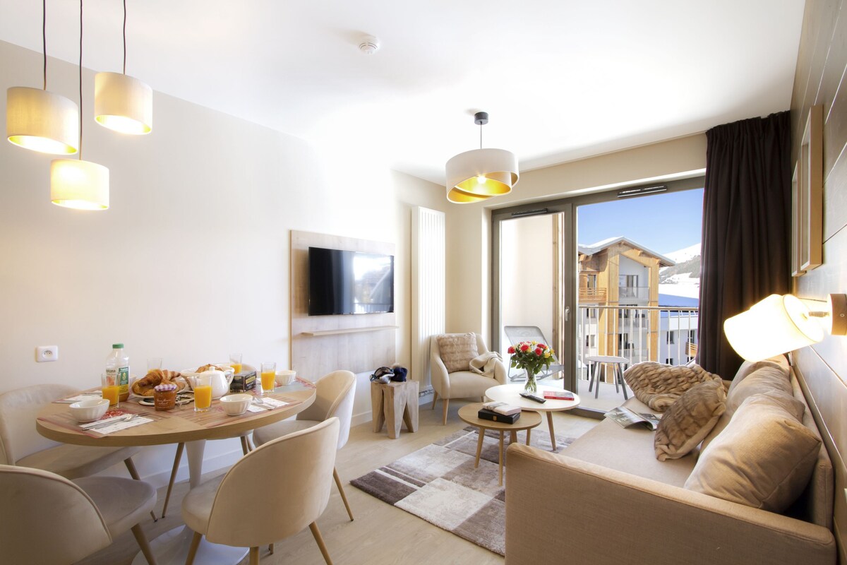 A modern hotel room with a dining area, a sofa, a TV, and a balcony with a view of buildings.