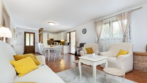 Un vaste salon avec un canapé blanc, des coussins jaunes, une table basse blanche et un fauteuil beige. On aperçoit une cuisine à l’arrière-plan, avec des meubles blancs et un coin repas.