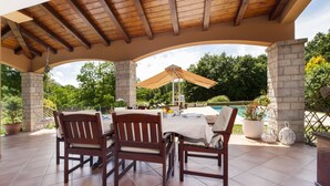 Une terrasse extérieure avec une table et des chaises en bois, un pilier en pierre et une vue sur une piscine.