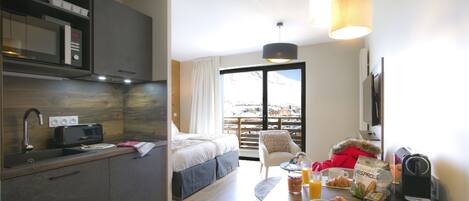 A modern hotel room with a kitchenette, a bed, a dining table with breakfast items, and a view of a snowy landscape.