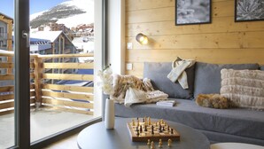 Et hyggeligt rum med trægulv, en grå sofa, et skakspil og udsigt til et snedækket bjerg.