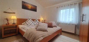 Un dormitorio con cama, lámparas de noche, un radiador, una ventana con cortinas y un cuadro colgado en la pared.