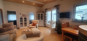 Una sala de estar acogedora con un sofá, un sillón y una mesa de comedor con sillas. Hay un aparador con puertas de vidrio y un televisor montado en la pared.