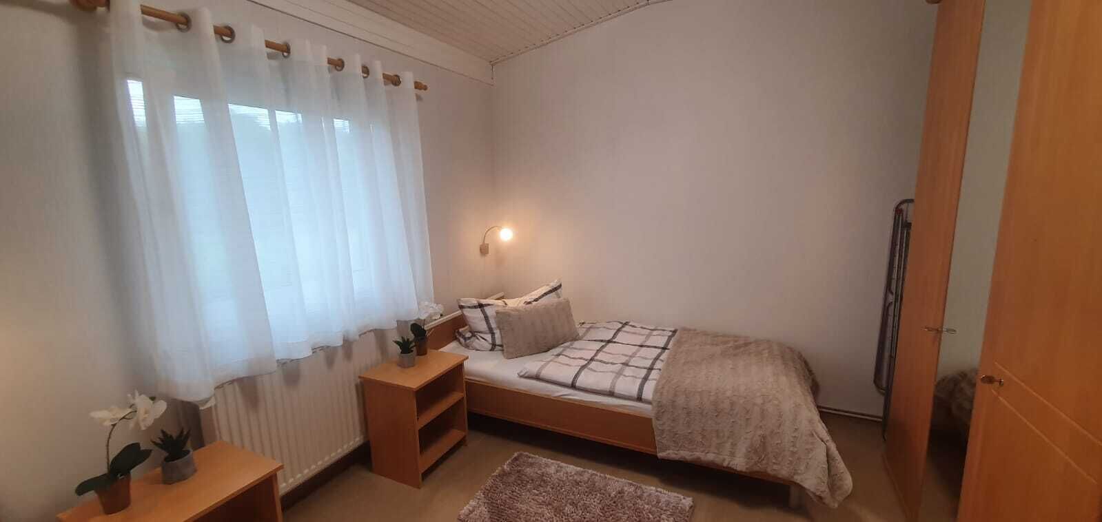 Een kleine, gezellige slaapkamer met een eenpersoonsbed, nachtkastje, radiator en raam met dunne gordijnen.