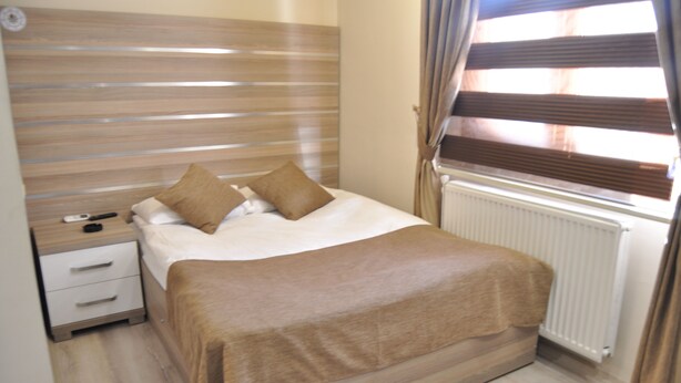 A neatly made bed with a brown bedspread and pillows, a bedside table with a lamp, and a window with blinds. - Arikan Otel (Pozanti)