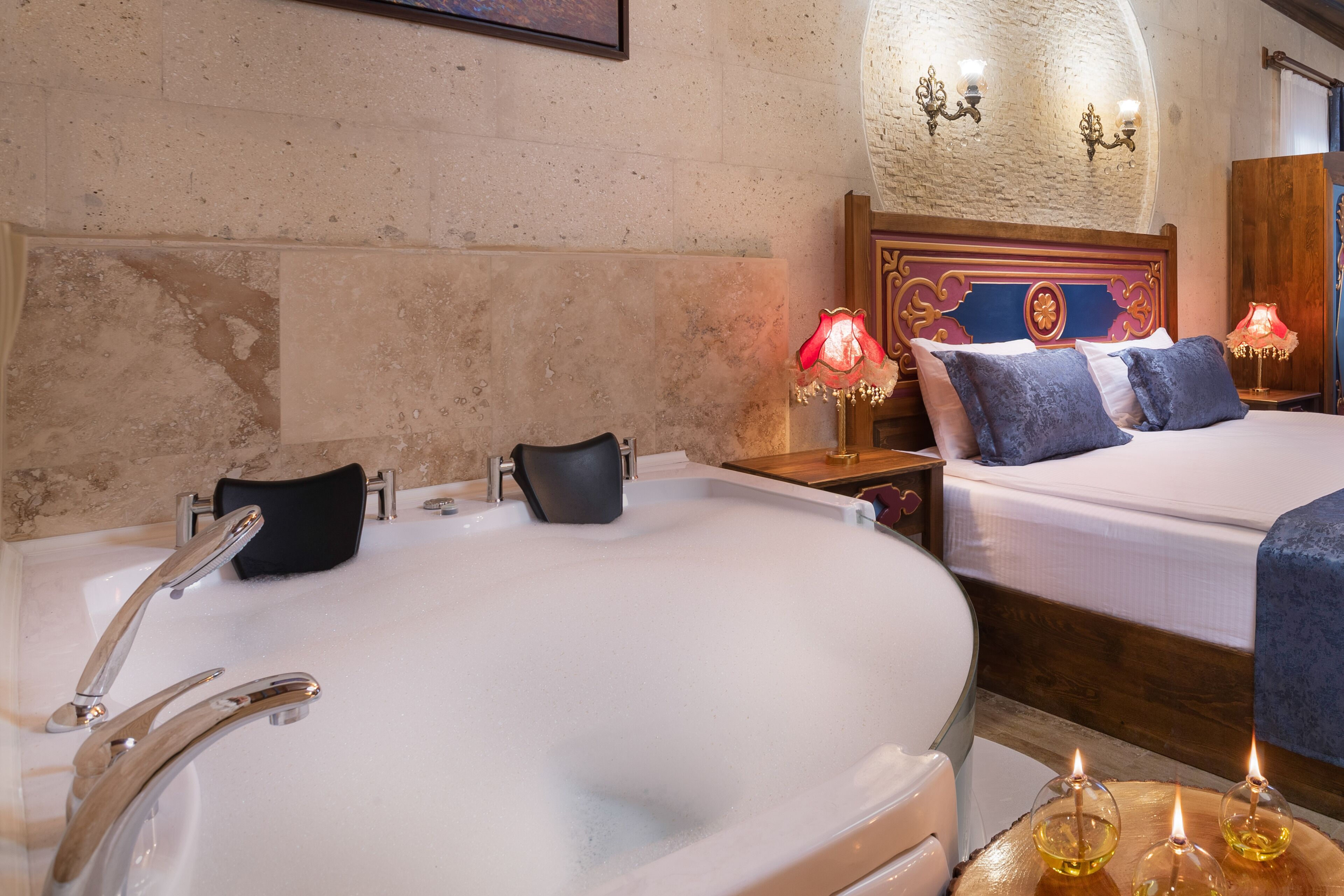 A hotel room with a large bathtub, two beds with blue bedding, a wooden headboard, and wall-mounted sconces.