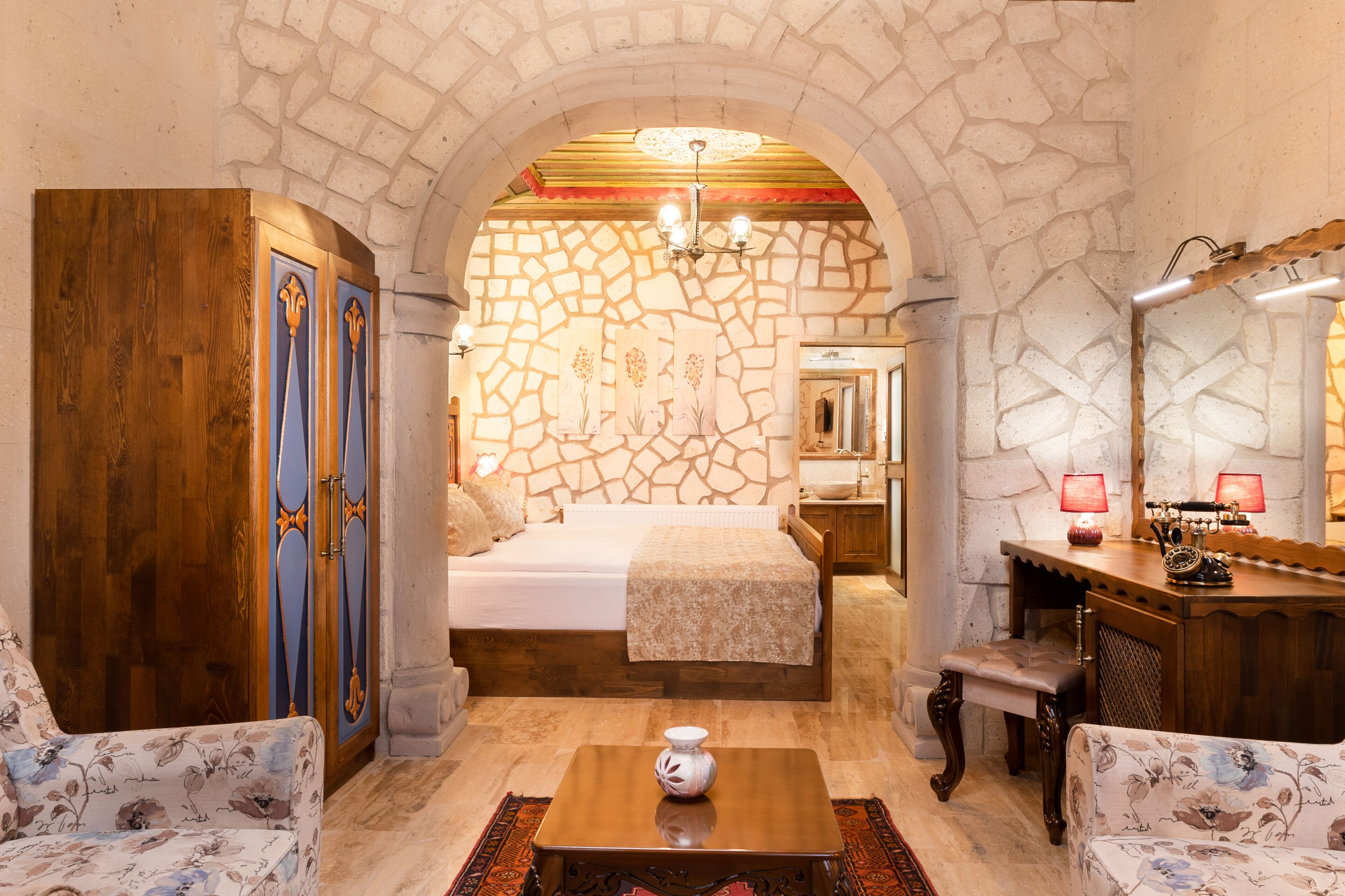 A room with a stone wall, a bed, a wooden wardrobe, a desk with a mirror, and two chairs.