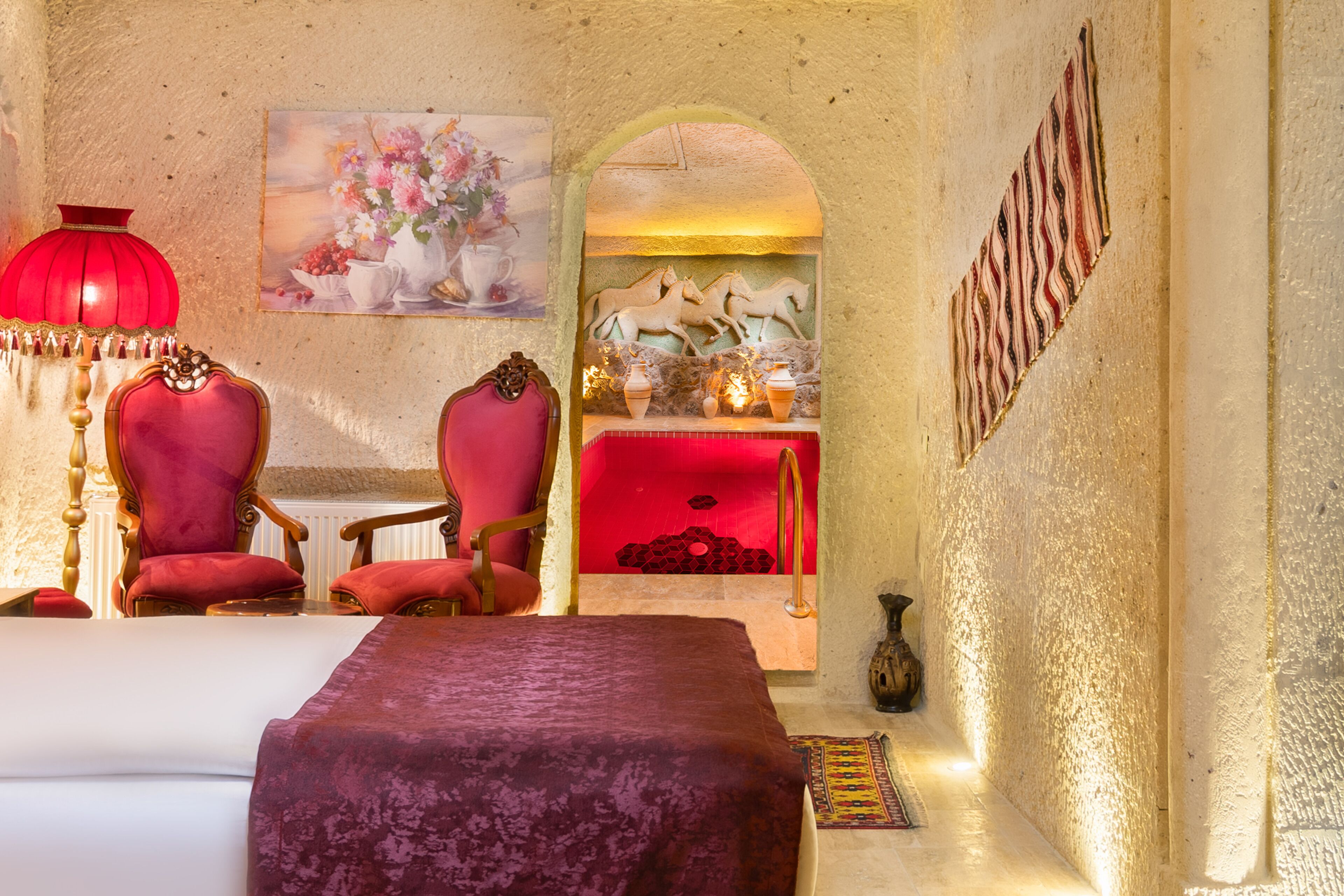 Una habitación con una cama, dos sillones rojos, una lámpara roja, un cuadro de flores, una chimenea con jarrones y una alfombra estampada.