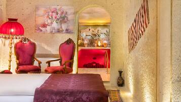 Una habitación con una cama, dos sillones rojos, una lámpara roja, un cuadro de flores, una chimenea con jarrones y una alfombra estampada.