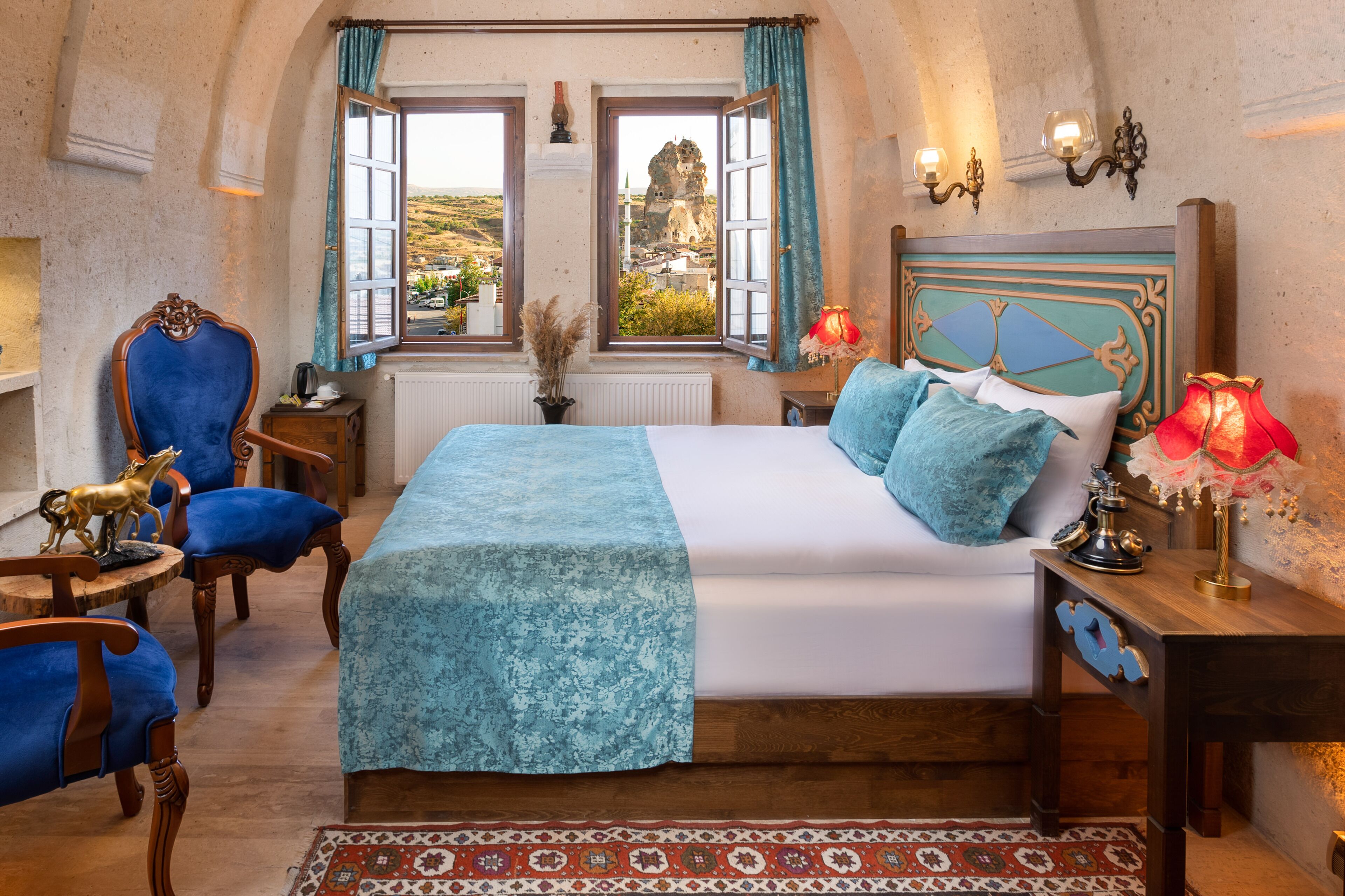 A bedroom with a large bed, two windows, a view of a rocky landscape, a wooden headboard, and a nightstand with a lamp.