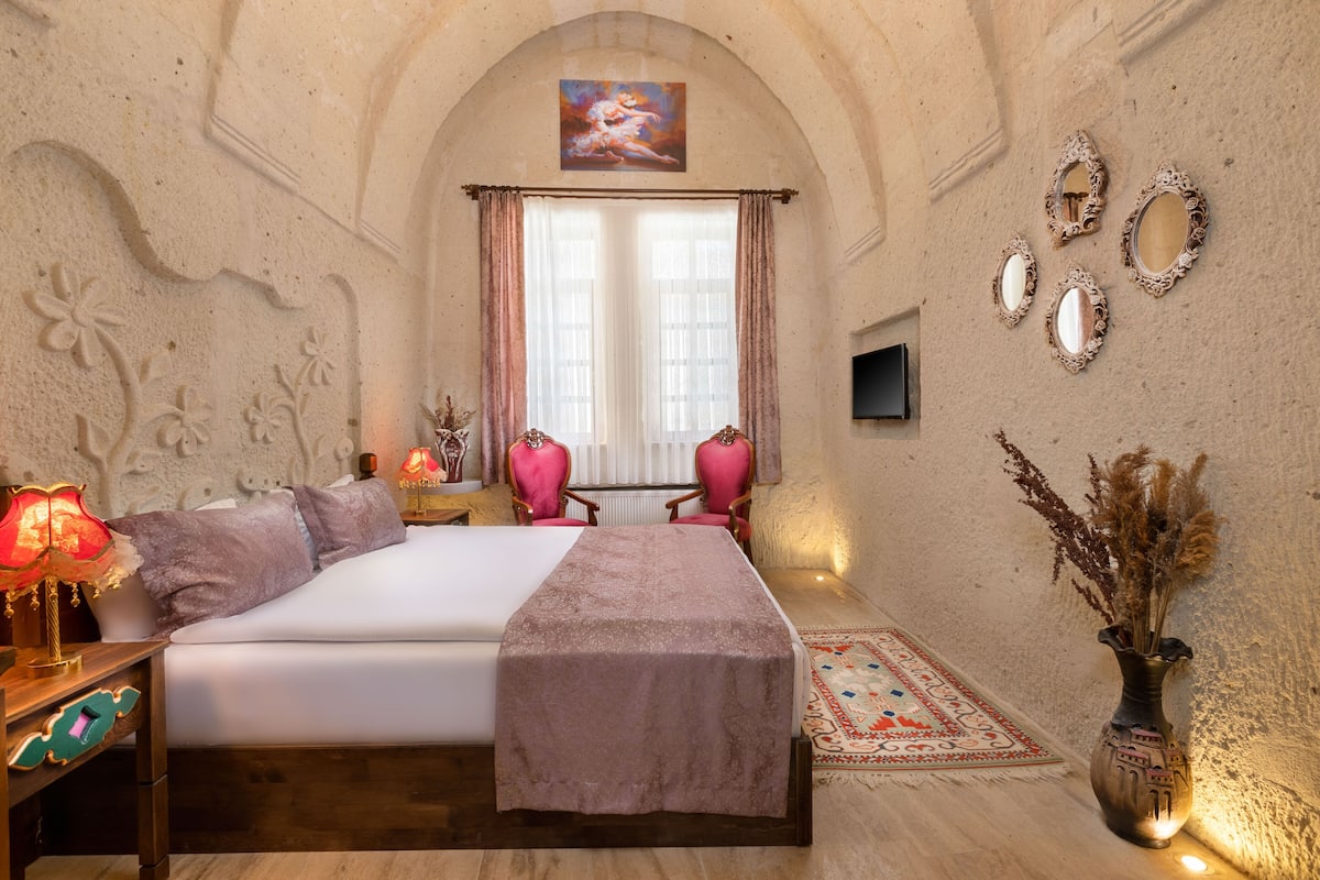 Un dormitorio con cama, dos sillas, una mesita de noche, una lámpara, un jarrón con plantas secas y espejos decorativos en la pared.