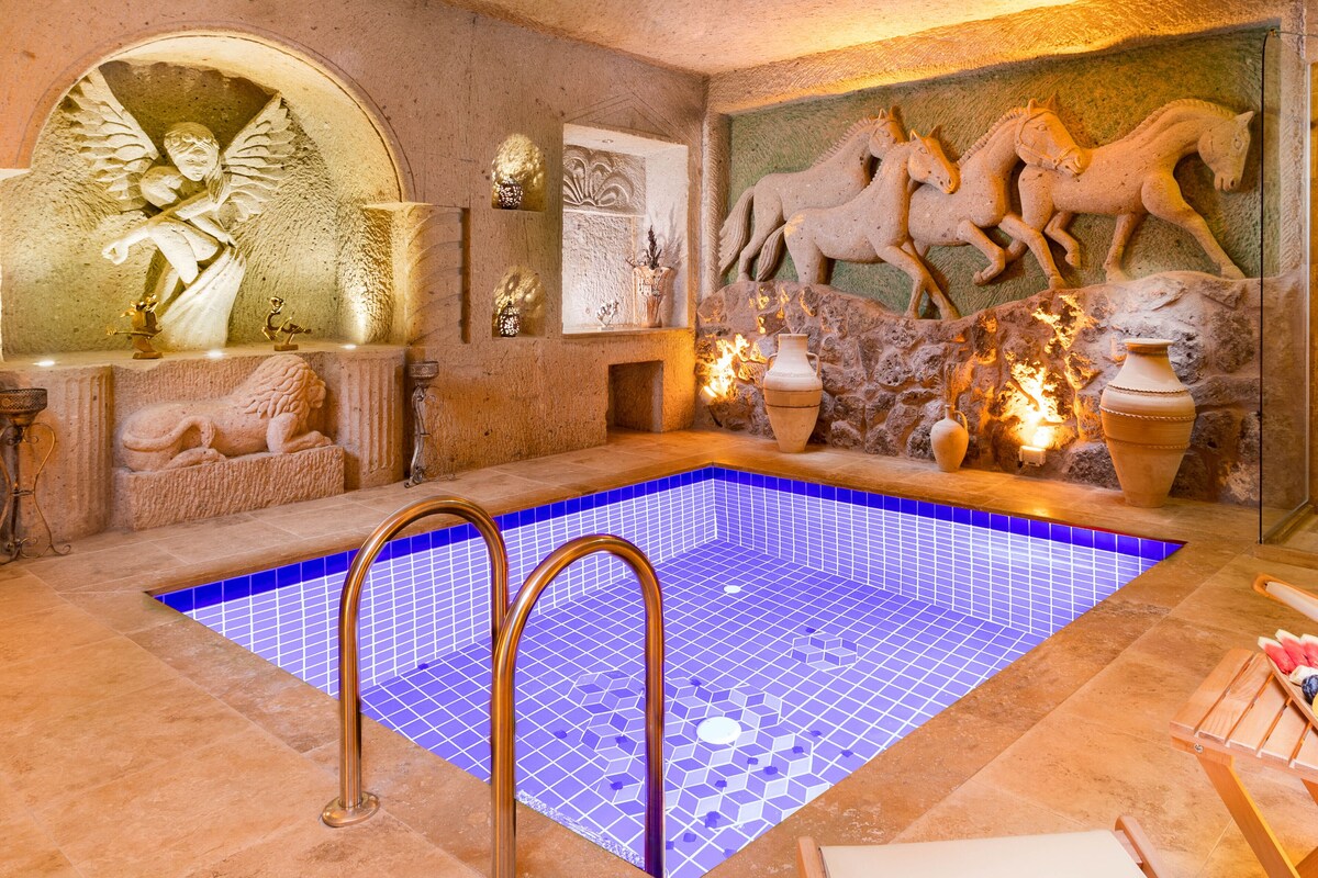 Uma área de piscina interior luxuosa com piso em mosaico, esculturas de pedra e uma parede de pedra com relevos de cavalos.