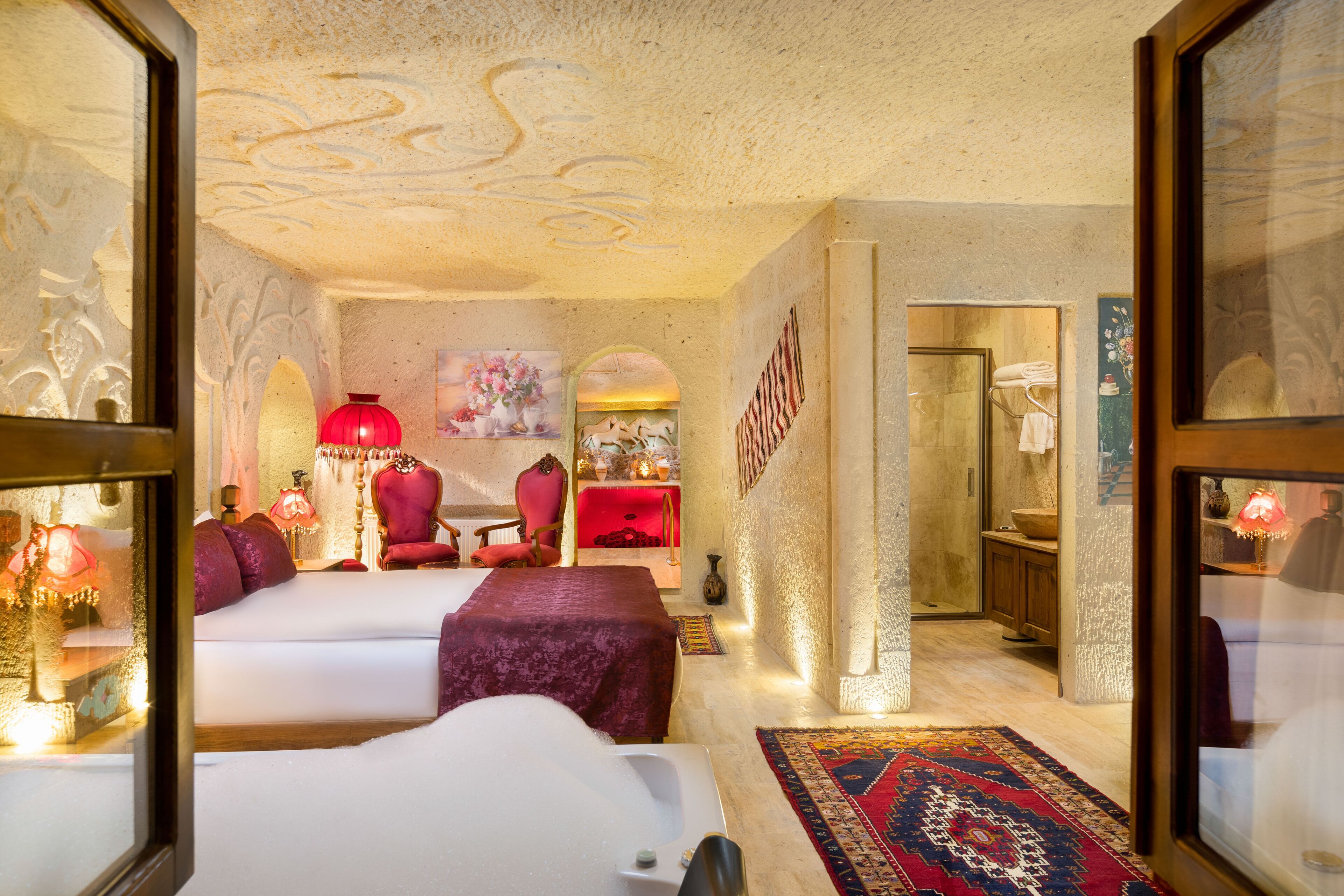 A hotel room with a large bed, a sitting area with a red chair, a small table, and a decorative rug.