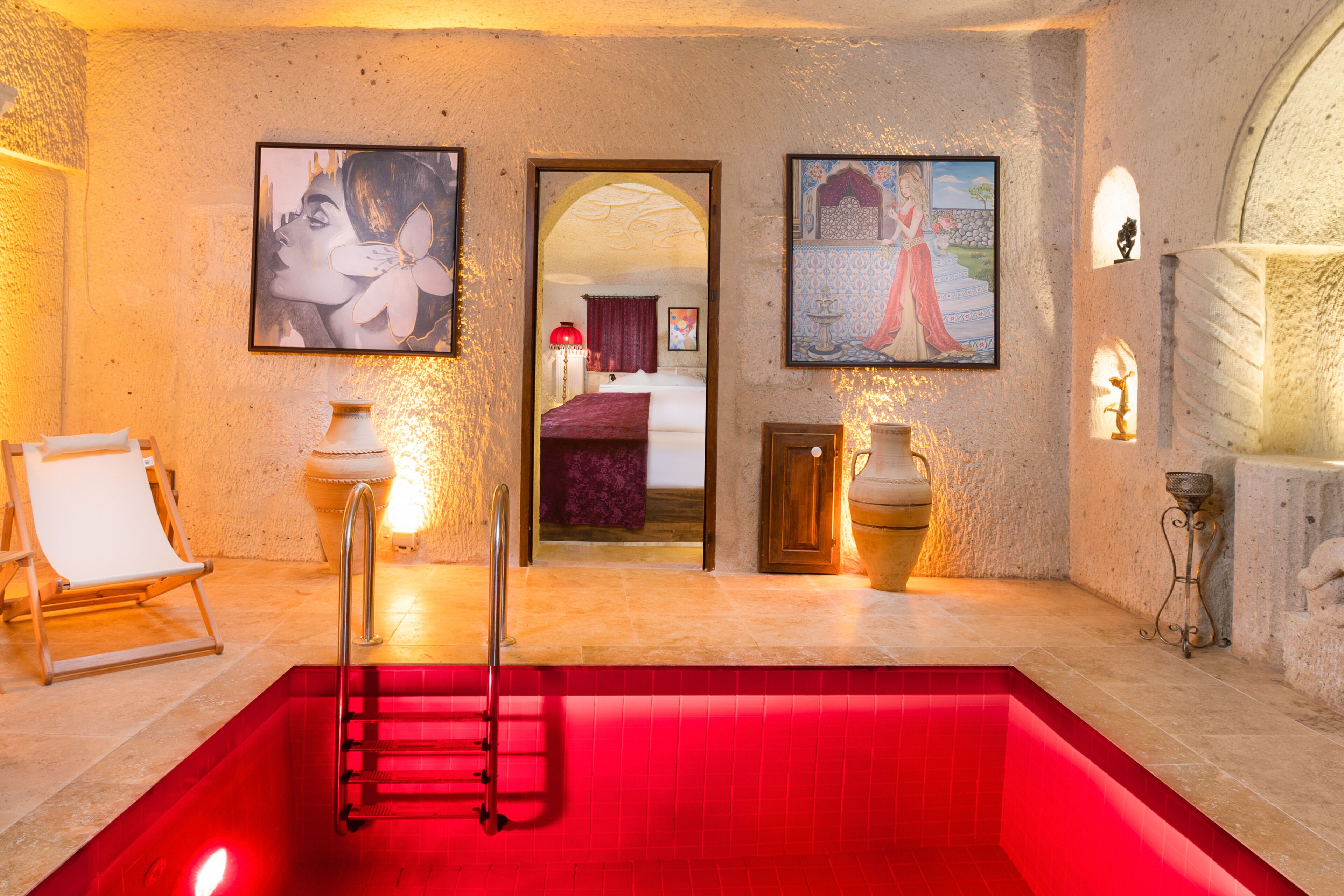A red heated pool with a ladder, framed artwork on the walls, and a view into a bedroom through an archway.