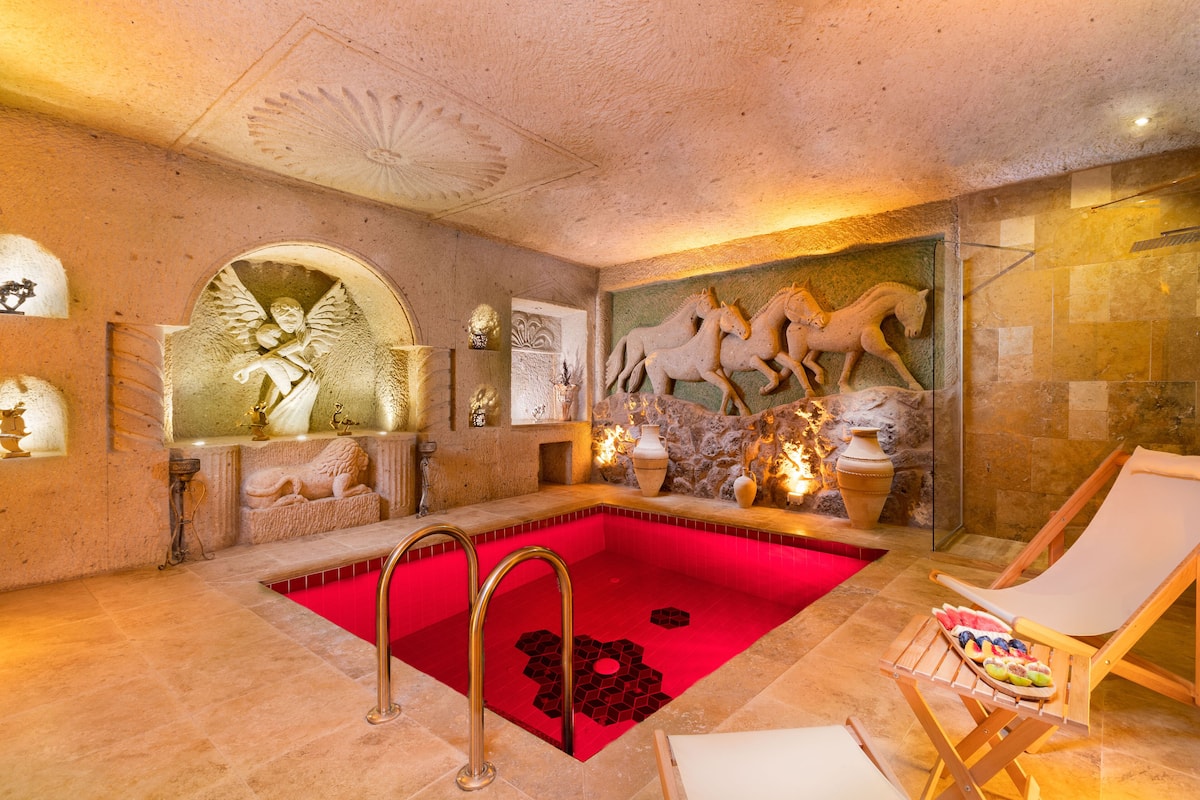 Un área de spa lujosa con piscina climatizada roja, una silla de lounge de madera y detalles artísticos en la pared, incluyendo esculturas de caballos.