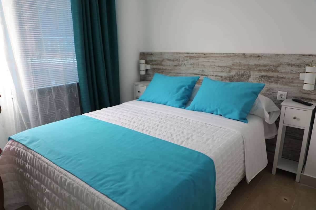 A neatly made bed with a white and blue bedspread, two matching pillows, and a wooden headboard. There are two bedside tables with lamps, and a window with curtains.