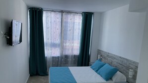 A bedroom with a bed, a nightstand, a TV, and a window with curtains. - Alojamiento Med The Blue Med. (El Campello)