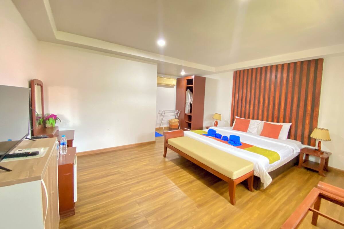 A hotel room with a wooden floor, a bed with colorful bedding, a desk with a computer, a wardrobe, and a mirror.