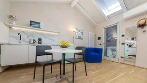 Une cuisine moderne avec un plan de travail blanc, un four à micro-ondes et une bouilloire. On y trouve une table ronde avec deux chaises et un vase rempli de fleurs jaunes. Une chaise bleue est placée à côté de la table.