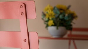 Une chaise rose avec une structure en métal et des vis, placée devant un arrière-plan flou où l’on aperçoit une plante en pot.