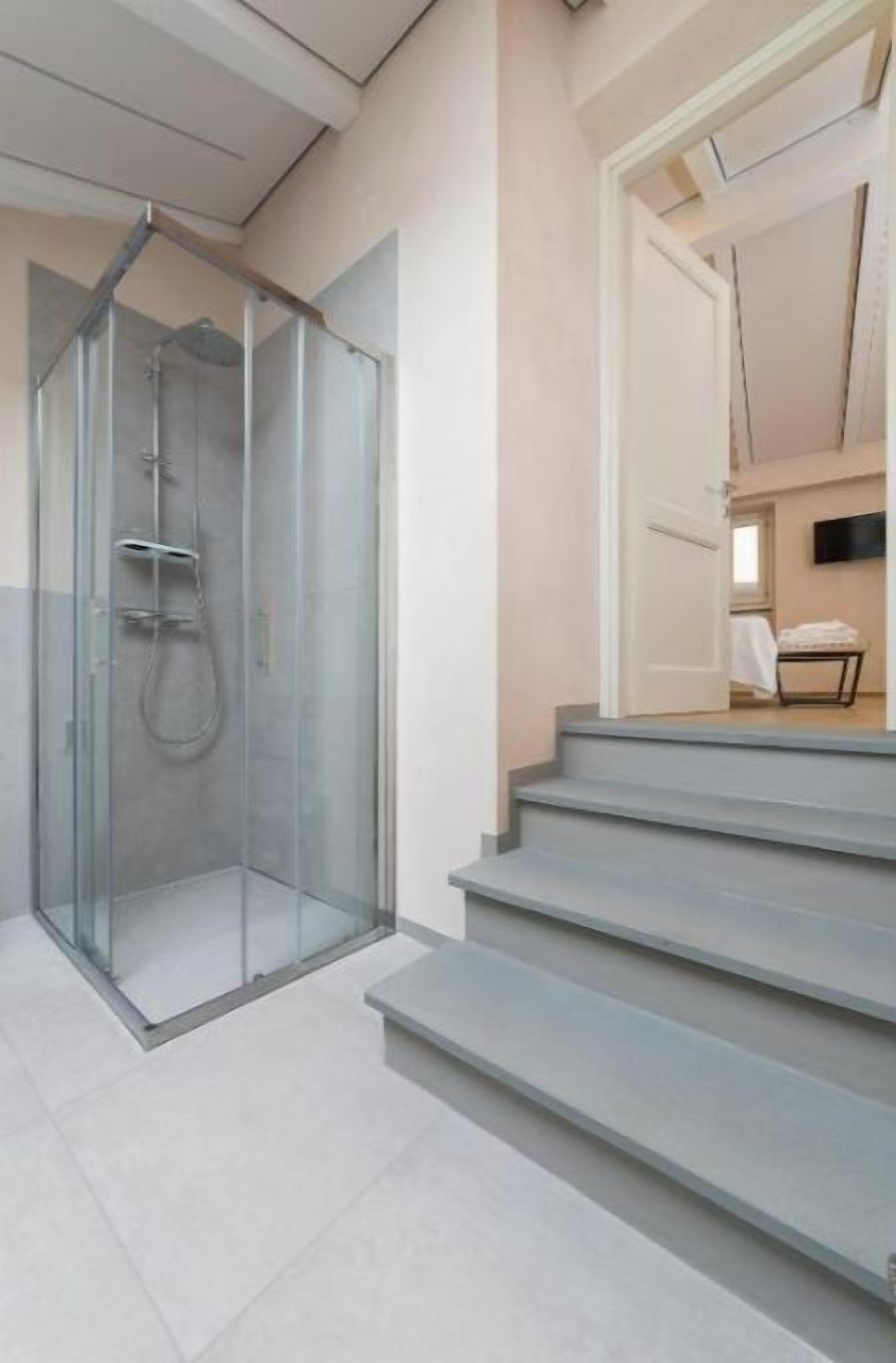 Un bagno moderno con doccia vetro, lavandino e WC. Sono presenti gradini che conducono a una porta che si apre su una zona giorno con televisore a schermo piatto.