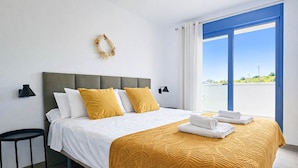 Una cama bien hecha con colcha blanca y mostaza, una mesita auxiliar y vistas al exterior a través de un ventanal grande.