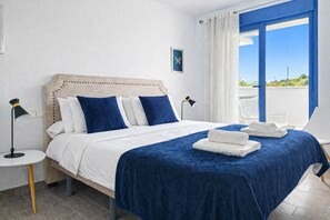 Un lit avec une tête de lit, une literie blanche et bleue, deux lampes de chevet et une vue sur l’extérieur par une fenêtre.
