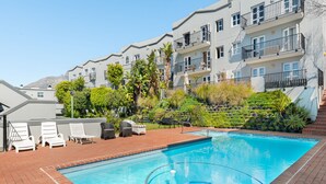A swimming pool with lounge chairs and a brick patio, surrounded by lush greenery and residential buildings. - De Waterkant Piazza 301 (Cape Town)
