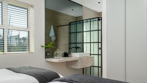 A modern bathroom with a glass shower enclosure, a built-in sink, and a mirror above it. There are two beds with gray blankets in the foreground. - De Waterkant Piazza 301 (Cape Town)