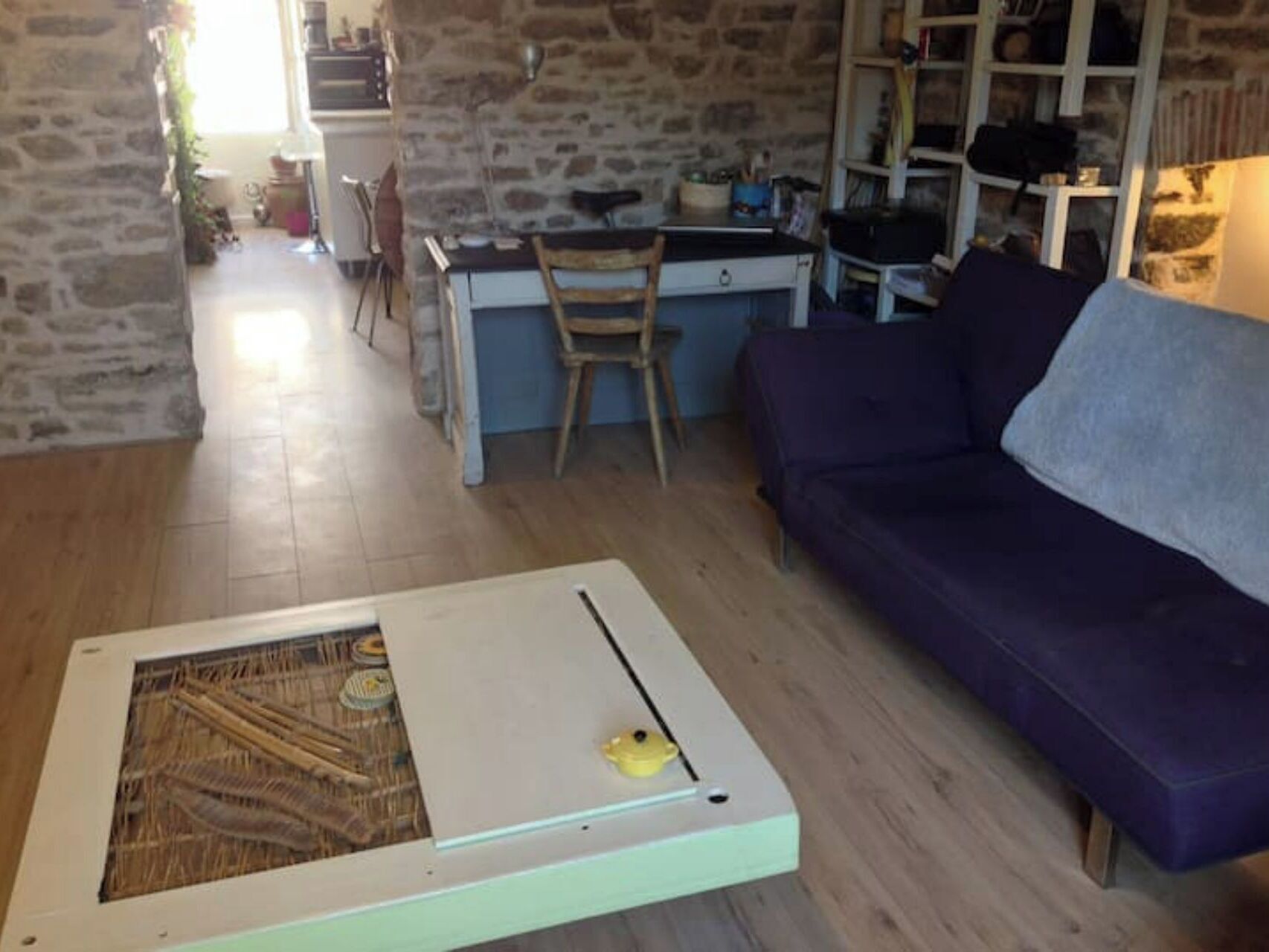 Un salon avec un canapé violet, une petite table sur laquelle est posé un cadre photo et un mur en pierre.