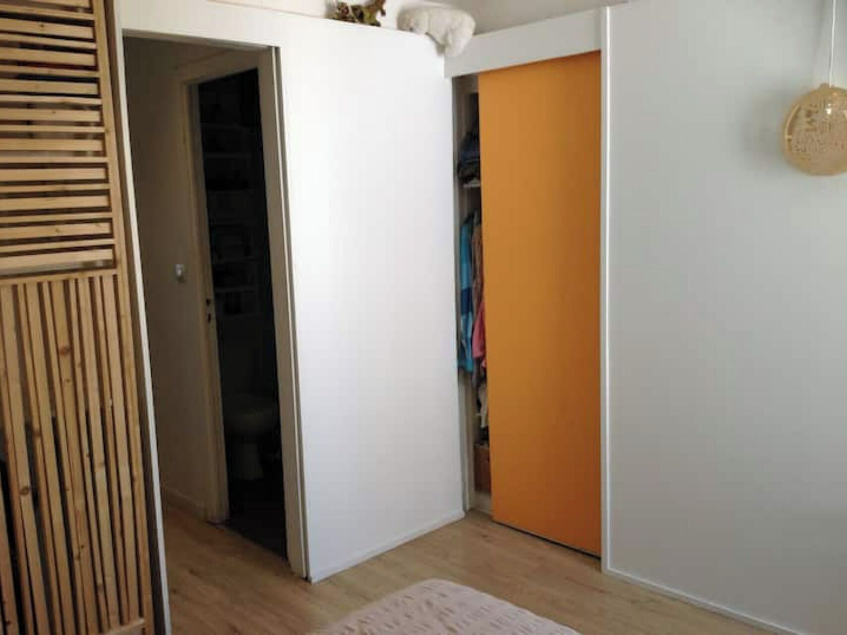 A room with a wooden slatted door, a white wardrobe, and an orange door leading to a closet with hanging clothes.