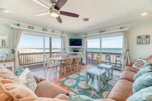 A spacious living room with a large window overlooking the beach, a ceiling fan, and a dining area with wooden furniture.