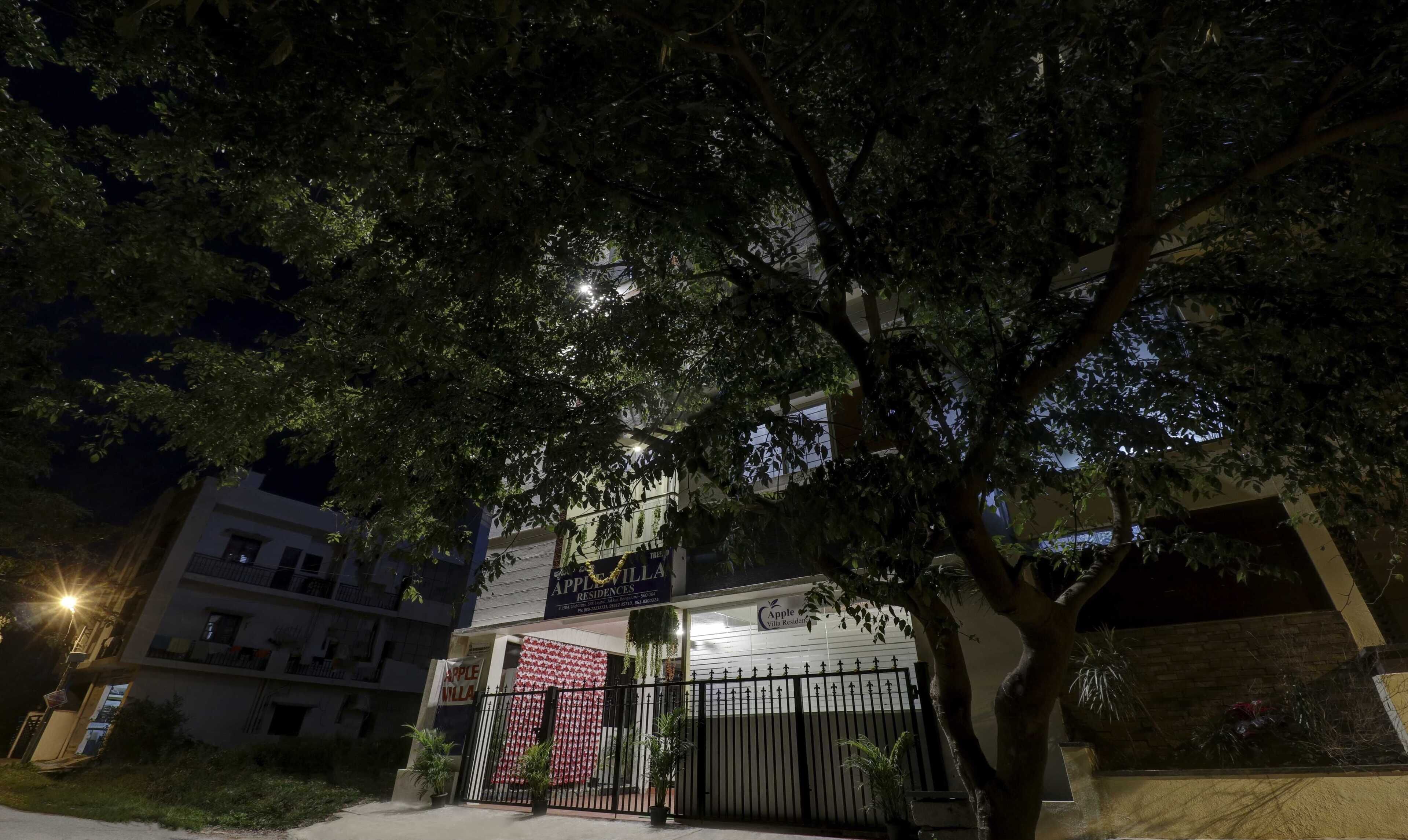 A gated residential complex with a large tree in the foreground, a well-lit entrance, and a sign for 'Apple Villa Residences'.