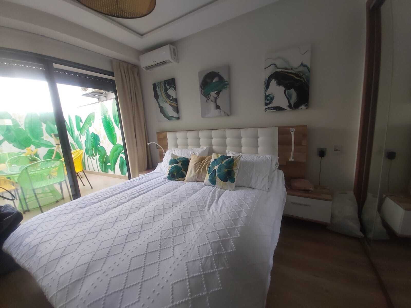 A bedroom with a large bed, a window with a view of greenery, and artwork on the wall.