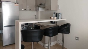 A compact kitchen with a refrigerator, a built-in oven, a microwave, and a coffee machine. There are three black bar stools and a white countertop.