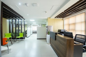 Zona de recepción con un mostrador de madera, una computadora e impresora. Hay sillas y una planta en maceta.
