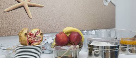 Une table sur laquelle se trouve un panier de fruits contenant des pommes, des bananes et une grenade, ainsi que plusieurs récipients et assiettes en verre.