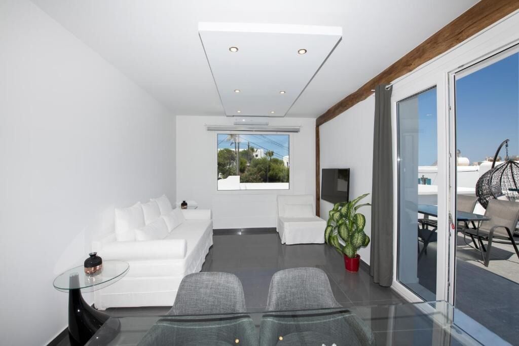 A modern living room with a white sofa, a glass coffee table, and a large window offering a view of the outdoors.